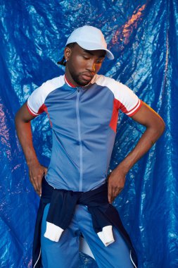 Modayı takip eden atletik kıyafetli kendine güvenen genç bir adam mavi bir zemine karşı duruyor..