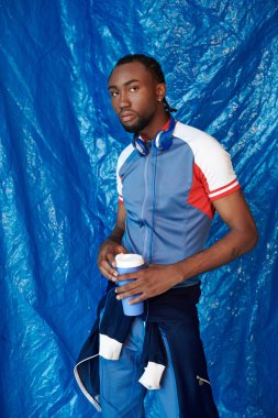 Kendine güvenen, şık bir atletizm kıyafeti giymiş genç bir adam mavi arka planda bir bardak tutuyor..