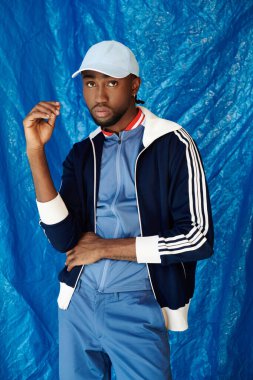 Yakışıklı genç bir adam kendine güvenerek poz veriyor, cesur mavi bir fon önünde modern bir kıyafet sergiliyor..