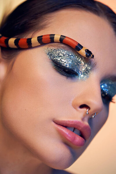 A young beautiful woman showcases a striking look with a colorful snake artfully draped on her brow.