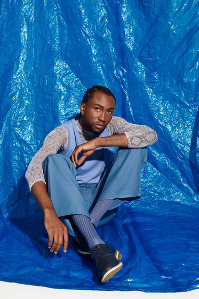 A charming young man in stylish attire sits gracefully against a blue backdrop.