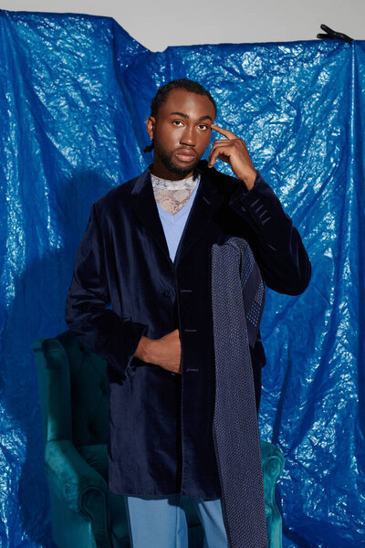 Молодой человек с яркими чертами лица уверенно стоит на ярком синем фоне.