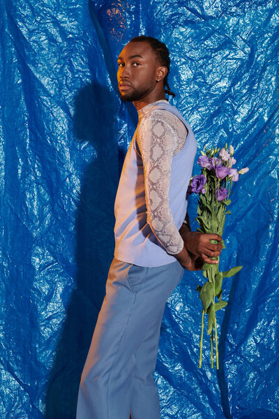 A handsome young man stands poised and stylish, holding a bouquet of flowers, radiating charm.