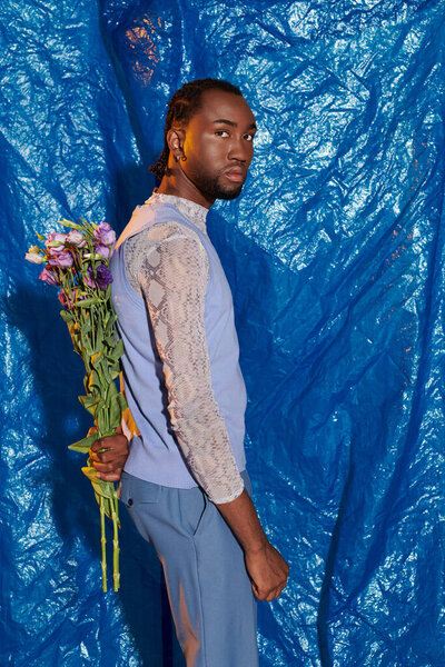 A stylish young man holds flowers behind his back against a bright blue background.