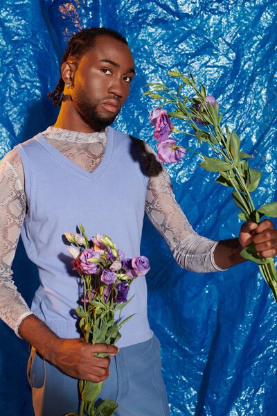 A stylish young man poses confidently, showcasing beautiful flowers in a vivid blue setting.