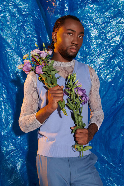 A confident young man in a stylish outfit holds purple flowers against a blue backdrop.