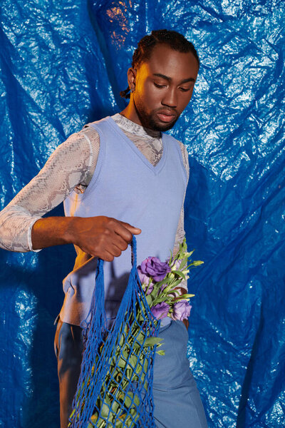 Handsome young man elegantly holds a bag with flowers while posing against a vivid blue backdrop.