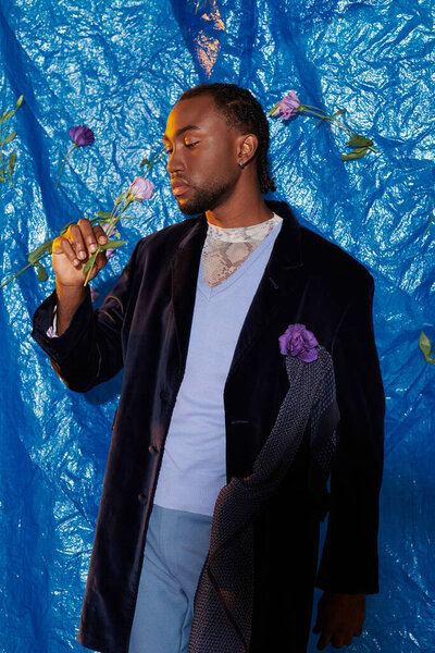 A stylish young man interacts with flowers against a striking blue backdrop.