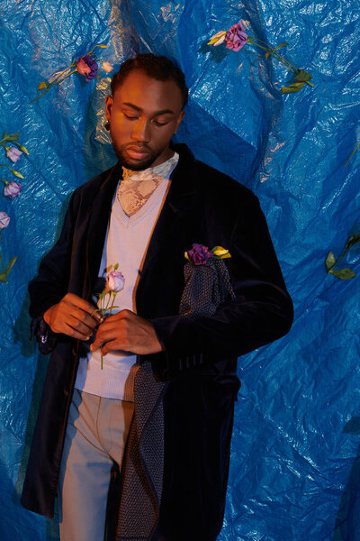 A young man poses in a stylish outfit against a bright blue background with floral accents.