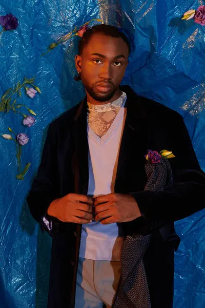 Yakışıklı genç bir adam koyu renk bir ceket ve açık renk süveter içinde dikiliyor, etrafı çiçeklerle çevrili..