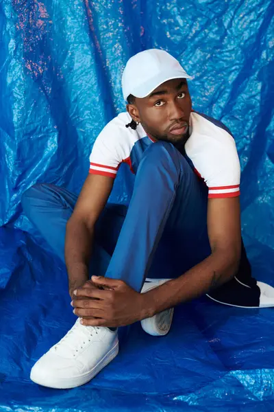 Düşünceli bir ifadeye sahip genç bir adam mavi bir arka planla çevrili yerde oturuyor.