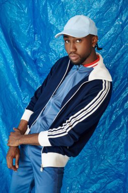 Şık giyinmiş yakışıklı genç bir adam canlı mavi bir arkaplan önünde güven içinde poz verir..