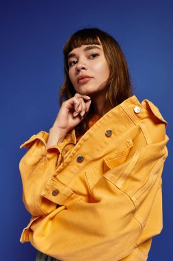 Kendine güvenen genç bir kadın, modaya uygun kıyafetlerini mavi arka planda çarpıcı bir pozla sergiliyor..