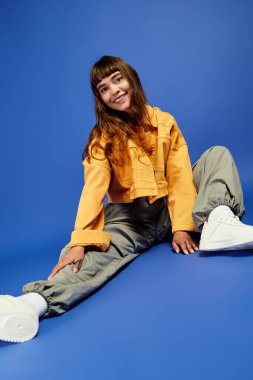 Genç bir kadın şık bir şekilde yerde oturuyor, parlak bir kıyafet giyiyor ve kendine güvenerek gülümsüyor..