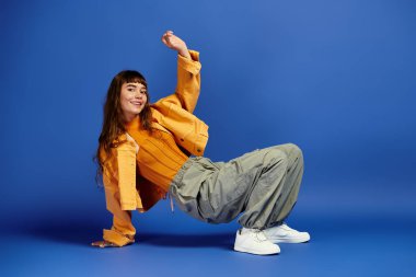 Genç bir kadın, mavi bir arka plana karşı neşeli bir pozla moda anlayışını sergiliyor..