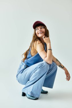 Moda kıyafetleri giymiş genç bir kadın, kapalı alandaki minimalist bir mekanda neşeli bir poz veriyor..