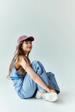 Şık genç bir kadın yerde oturuyor, kot pantolon ve kep giymiş, kendinden çok emin..