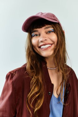 Moda kıyafetleri içinde kendine güvenen genç bir kadın geniş bir gülümseme sergiliyor, eşsiz bir şapka takıyor ve cesur bir makyaj yapıyor..