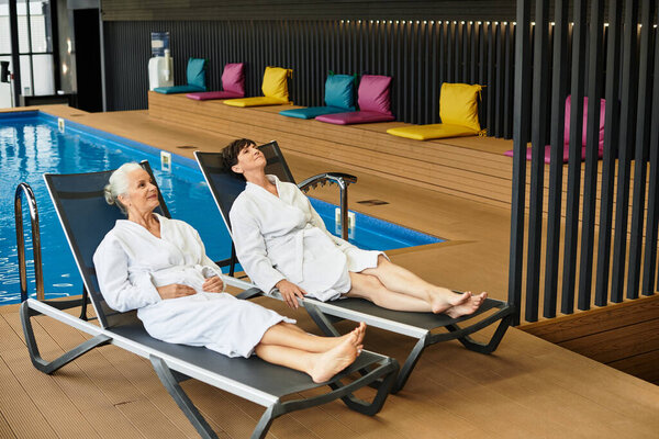 Two beautiful mature women unwind in spa robes beside a serene pool area, embracing tranquility.