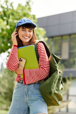 Güzel kız öğrenci elinde defterlerle dışarıda duruyor ve güneşin altında neşeyle gülümsüyor..