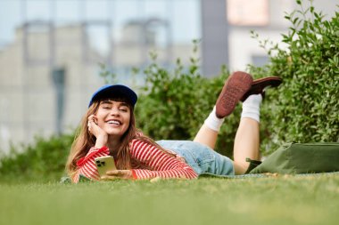 Neşeli bir öğrenci çimenlerde dinlenir, telefonuyla meşgul olur ve sıcak güneş ışığının tadını çıkarır..