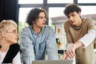 Üç genç profesyonel dikkatle dizüstü bilgisayara odaklanarak parlak bir ofisin ortasında beyin fırtınası yapıyorlar..