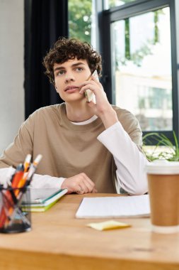 Ofis malzemeleriyle çevrili genç bir adam telefonda konuşuyor..