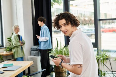 Kıvırcık saçlı genç bir adam akıllı telefonunu kullanırken meslektaşları parlak bir ofiste sohbet ediyorlar..