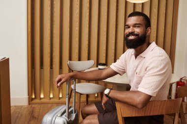 Sakallı, gülümseyen bir adam şık bir otel lobisinde rahatça oturur ve gezisine hazırlanır..