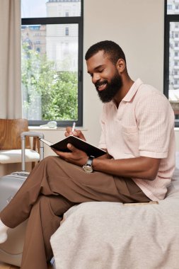 Modern bir otel odasında dinlenen, bakımlı bir adam, bir deftere yazarak seyahat sükunetini kucaklıyor..
