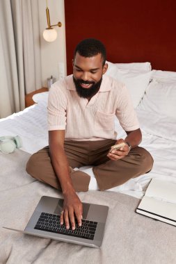 Çekici bir adam otel odasında rahatça oturuyor ve dizüstü bilgisayarı üzerinde çalışıyor..