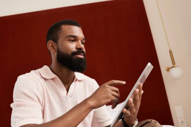 İyi bakımlı bir siyah adam modern bir otel odasında dinleniyor ve seyahatlerini tabletle planlıyor..