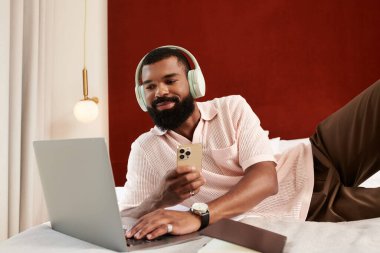 Zarif bir Afrikalı Amerikalı adam şık bir otel odasında telefonunu ve laptopunu kullanarak rahatlıyor..