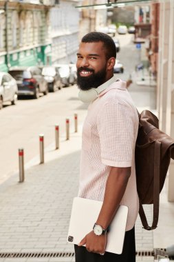 Zarif bir Afro-Amerikan erkeği sırt çantası taşırken gülümseyerek şehir macerasından zevk alır..