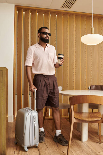 A dapper African American man stands in a modern hotel cafe with a suitcase and coffee.