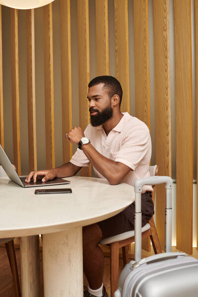 Стильный черный мужчина сидит за столом в современном вестибюле отеля, сосредоточенный на своем ноутбуке.