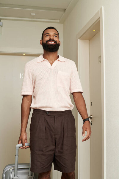 Стильный африканский американец входит в современный номер отеля с улыбкой после путешествия.
