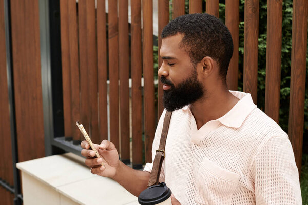 Стильный черный мужчина пьет кофе и проверяет телефон, наслаждаясь оживленным городом..