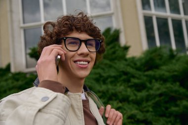 Açık havada telefonunda kıvırcık saçlı neşeli genç bir adam. Etrafı yeşilliklerle çevrili..