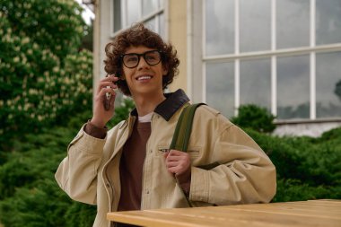 Neşeli genç bir adam dışarıda yürüyor, yeşil bir çevrede telefonla konuşuyor..