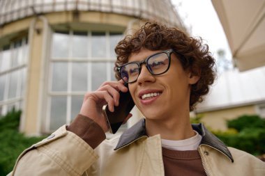 Kıvırcık saçlı ve gözlüklü genç bir adam dışarıda yürüyor, mutlu bir şekilde telefonla konuşuyor..