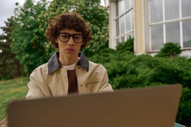 Kıvırcık saçlı ve gözlüklü genç bir öğrenci laptopunu kullanarak dışarı çıkıyor..
