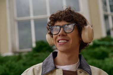 Yakışıklı bir öğrenci dışarıda kulaklıklarla sakince yürüyor, çevreyi izlerken gülümsüyor..