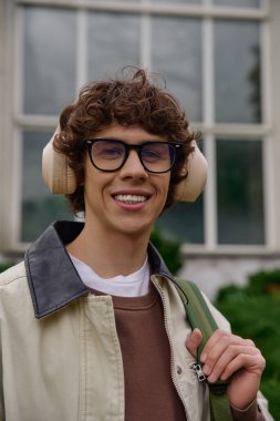 Kıvırcık saçlı yakışıklı öğrenci yeşil bir ortamda yürüyor, büyüleyici ve neşe saçıyor..