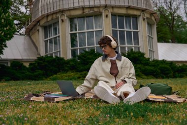 Çevresi yeşilliklerle çevriliyken, yakışıklı genç bir öğrenci dizüstü bilgisayarıyla çimlerin üzerinde oturuyor..