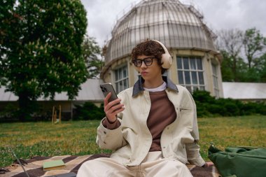 Yakışıklı genç bir öğrenci, yeşil bir ortamda telefonunu kullanarak battaniyenin üzerinde dinleniyor..