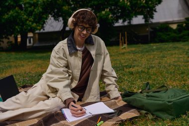 Genç adam parkta bir battaniyeye uzanıyor, notlar yazıyor ve güzel havanın tadını çıkarıyor..