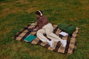 Genç, yakışıklı bir öğrenci dışarıda bir battaniyenin üzerinde rahatlarken dizüstü bilgisayarla ders çalışıyor..