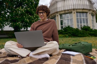 Genç bir adam, güzel bir açık hava ortamında laptopuyla rahatça bir battaniyeye oturur..