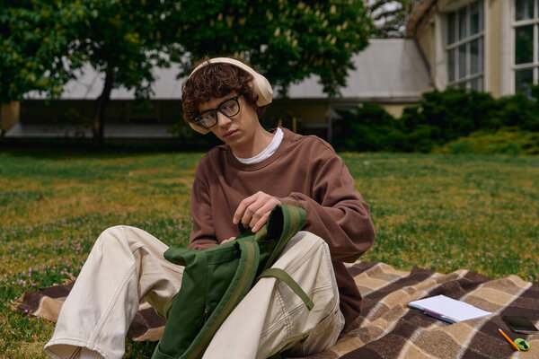 A young and handsome student takes a stroll outdoors, examining items in his backpack on soft grass.